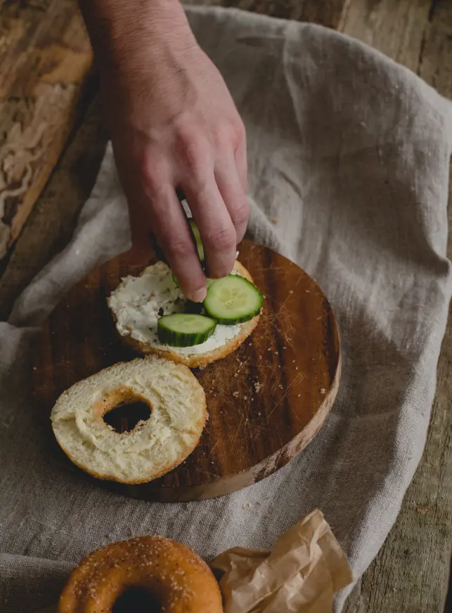 A hand placing cucumber slices on a bagel with cream cheese, set on a wooden board with a rustic background.