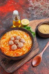 A bowl of pasta meatball soup topped with tender meatballs, served on a wooden board with a side of uncooked pasta, olive oil, lemon, and fresh parsley.