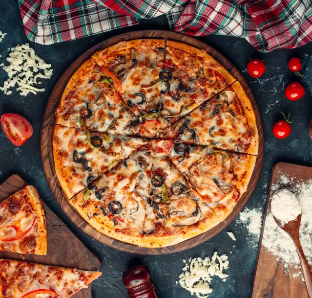 A freshly baked pizza with a variety of toppings, including olives, mushrooms, tomatoes, and melted cheese, served on a wooden board with scattered ingredients like shredded cheese and cherry tomatoes.