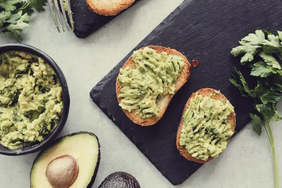 Two slices of toasted bread topped with mashed avocado, placed on a black slate board with a bowl of mashed avocado, fresh parsley, and whole avocados in the background.