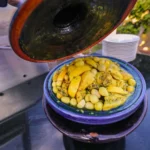 A close-up view of a traditional Moroccan chicken tagine, topped with preserved lemons and green olives, served in a blue ceramic tagine dish with its lid partially lifted.