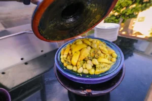 A close-up view of a traditional Moroccan chicken tagine, topped with preserved lemons and green olives, served in a blue ceramic tagine dish with its lid partially lifted.