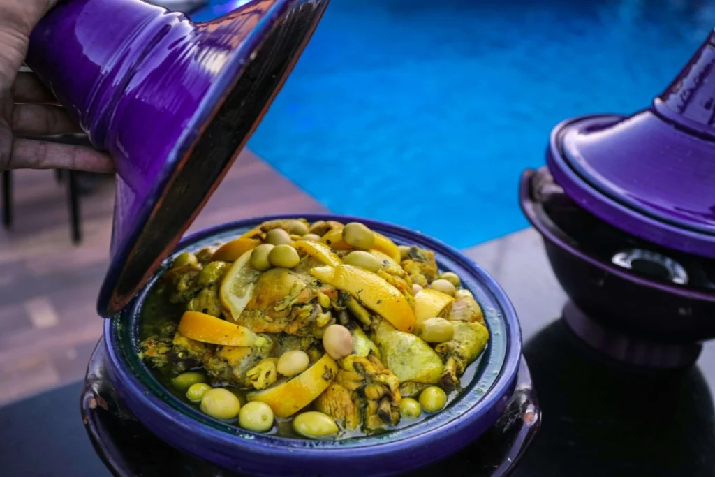 A vibrant purple tagine lid being lifted to reveal a beautifully cooked Moroccan chicken tagine with preserved lemons and green olives, set against a backdrop of a poolside setting.