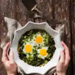 A white ceramic dish with a spinach and egg scramble topped with three sunny-side-up eggs, held by two hands over a rustic wooden table.