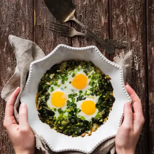 A white ceramic dish with a spinach and egg scramble topped with three sunny-side-up eggs, held by two hands over a rustic wooden table.