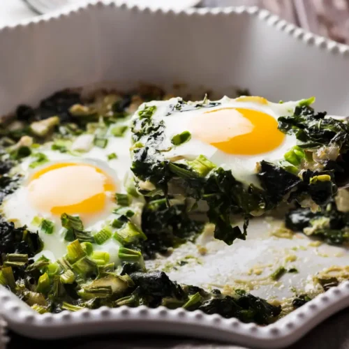 Close-up of a spinach and egg scramble topped with sunny-side-up eggs, garnished with chopped green onions, served in a white dish.