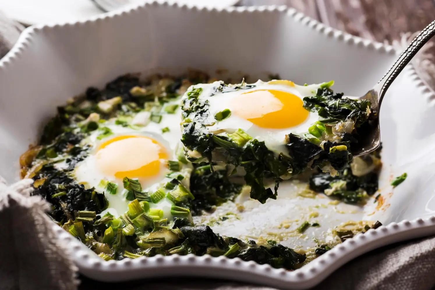 Close-up of a spinach and egg scramble topped with sunny-side-up eggs, garnished with chopped green onions, served in a white dish.