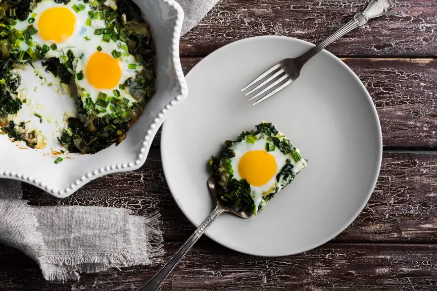 Spinach and egg scramble in a white ceramic dish, with a serving on a plate topped with a sunny-side-up egg, placed on a rustic wooden table.