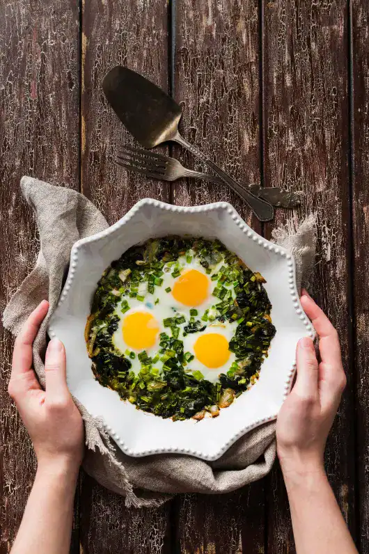 A white ceramic dish with a spinach and egg scramble topped with three sunny-side-up eggs, held by two hands over a rustic wooden table.