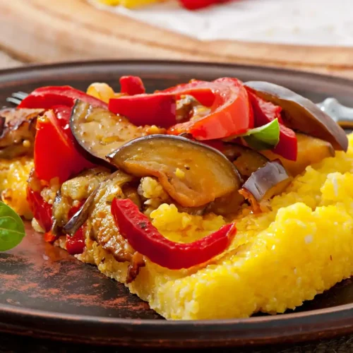 A slice of golden polenta topped with roasted eggplant, red peppers, and fresh basil leaves, served on a rustic brown plate.