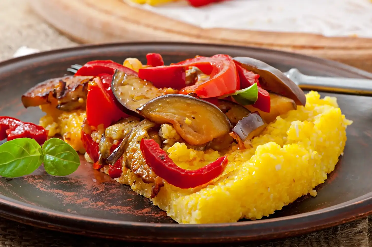 A slice of golden polenta topped with roasted eggplant, red peppers, and fresh basil leaves, served on a rustic brown plate.