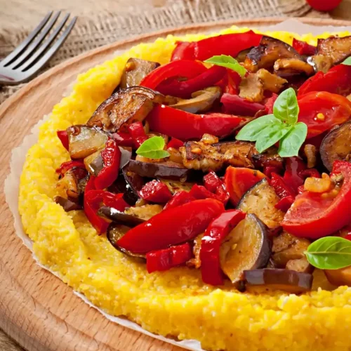 A round serving of creamy polenta topped with roasted eggplant, red peppers, cherry tomatoes, and fresh basil, displayed on a wooden platter.