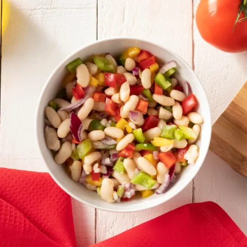 A colorful bowl of white bean salad with diced red and green bell peppers, yellow peppers, and red onions, set against a white wooden background with a red napkin, a tomato, and a sliced onion.