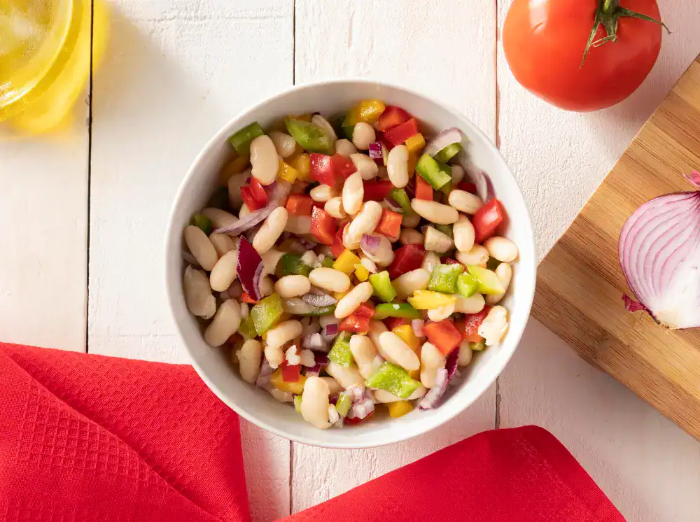 A colorful bowl of white bean salad with diced red and green bell peppers, yellow peppers, and red onions, set against a white wooden background with a red napkin, a tomato, and a sliced onion.