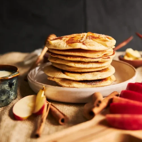 A stack of apple pancakes on a ceramic plate, accompanied by sliced red apples, cinnamon sticks, and a bowl of cream, set on a rustic table.