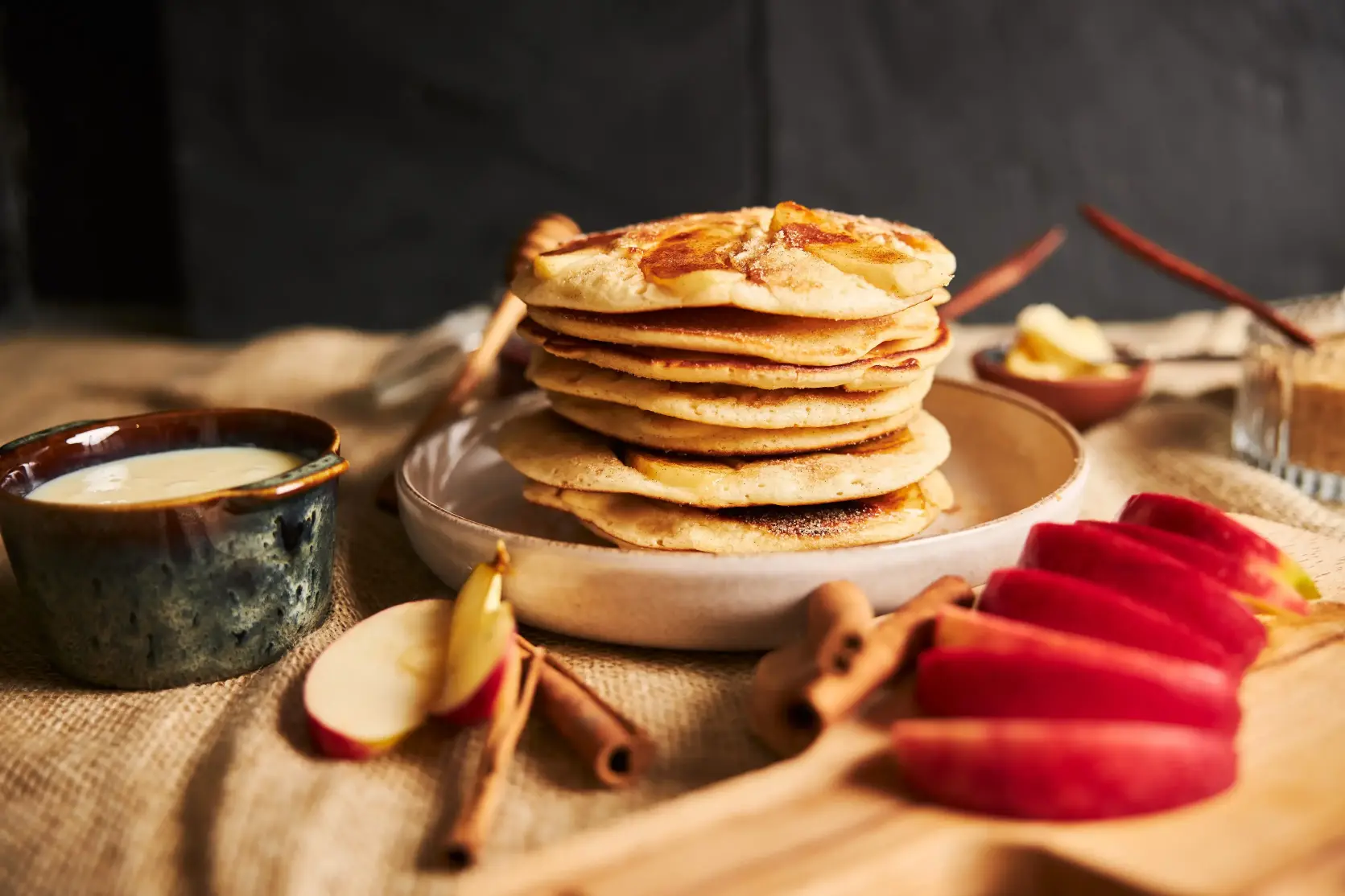 A stack of apple pancakes on a ceramic plate, accompanied by sliced red apples, cinnamon sticks, and a bowl of cream, set on a rustic table.