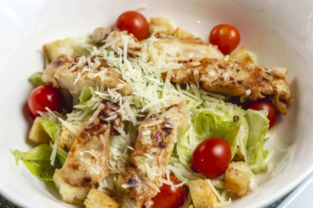 A classic chicken Caesar salad in a white bowl, featuring grilled chicken strips, shredded parmesan, crisp lettuce, cherry tomatoes, and croutons.