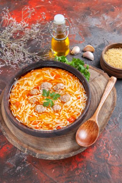 A bowl of hearty pasta meatball soup with noodles, garnished with fresh parsley, served on a wooden board with a wooden spoon, garlic, olive oil, and uncooked pasta nearby.