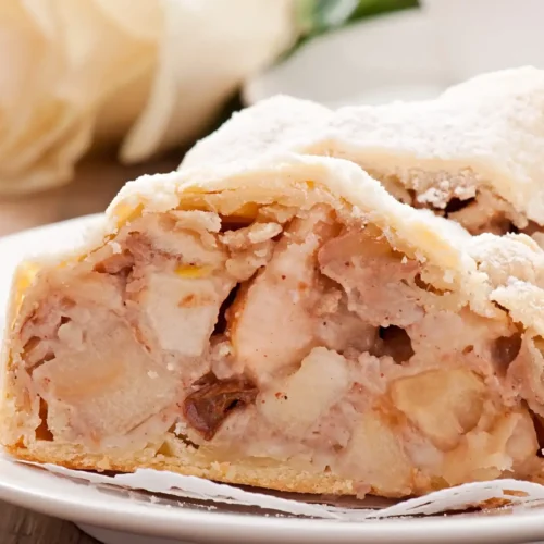 A close-up of a slice of apple strudel with a flaky crust and creamy, spiced apple filling, dusted with powdered sugar and served on a white plate.