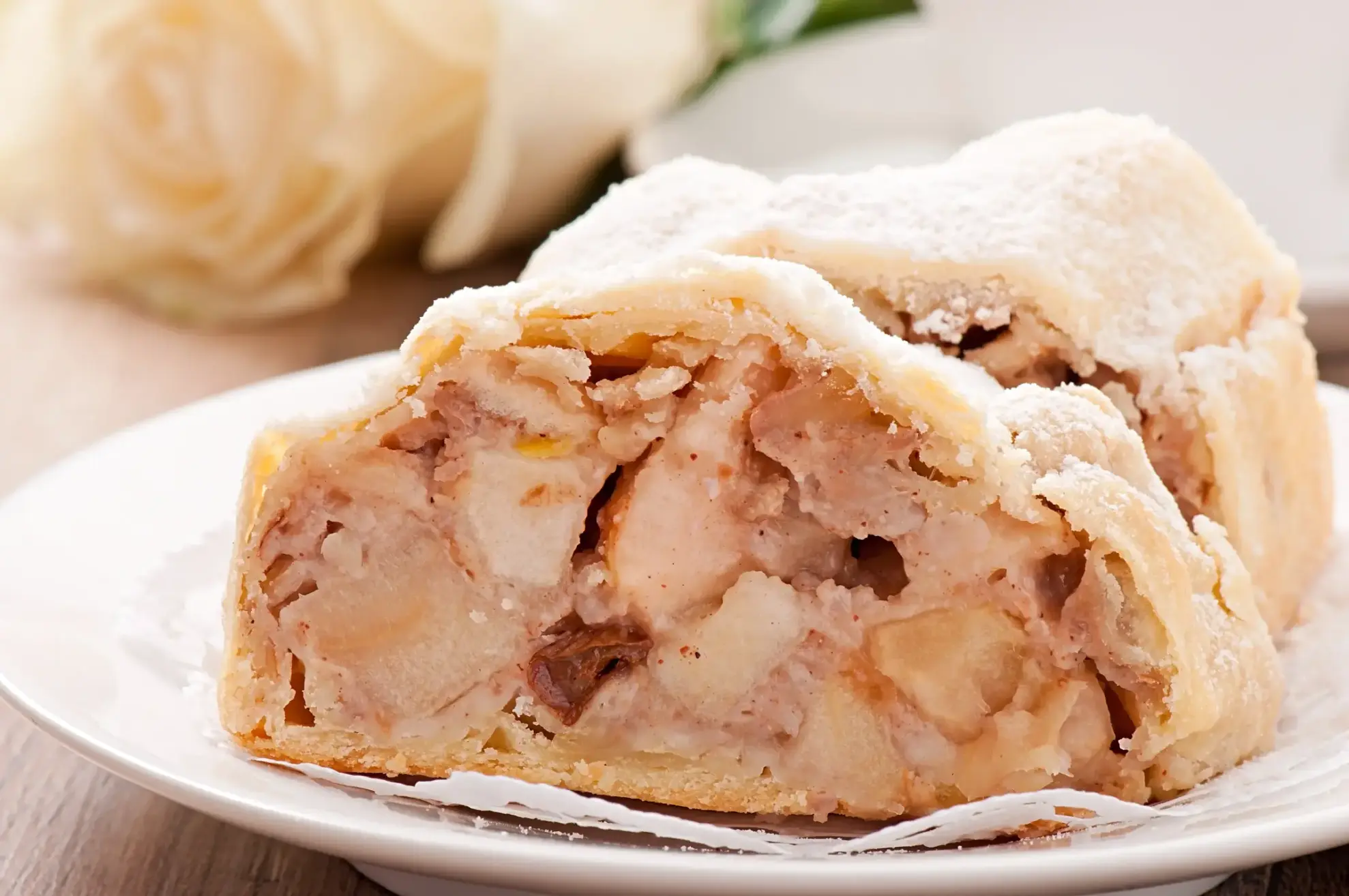 A close-up of a slice of apple strudel with a flaky crust and creamy, spiced apple filling, dusted with powdered sugar and served on a white plate.