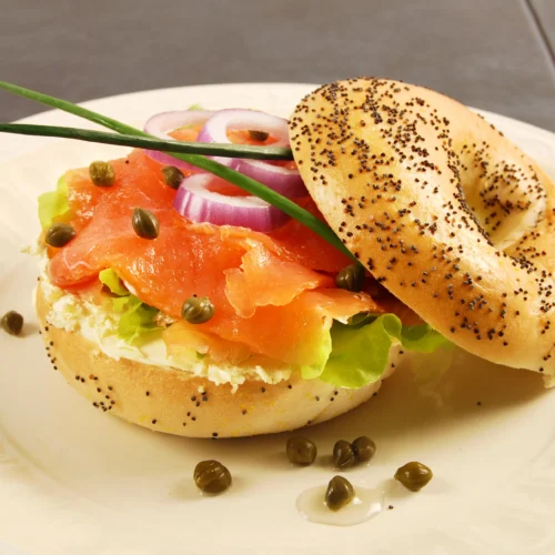 Poppy seed bagel filled with smoked salmon, cream cheese, capers, red onion, and lettuce on a white plate.