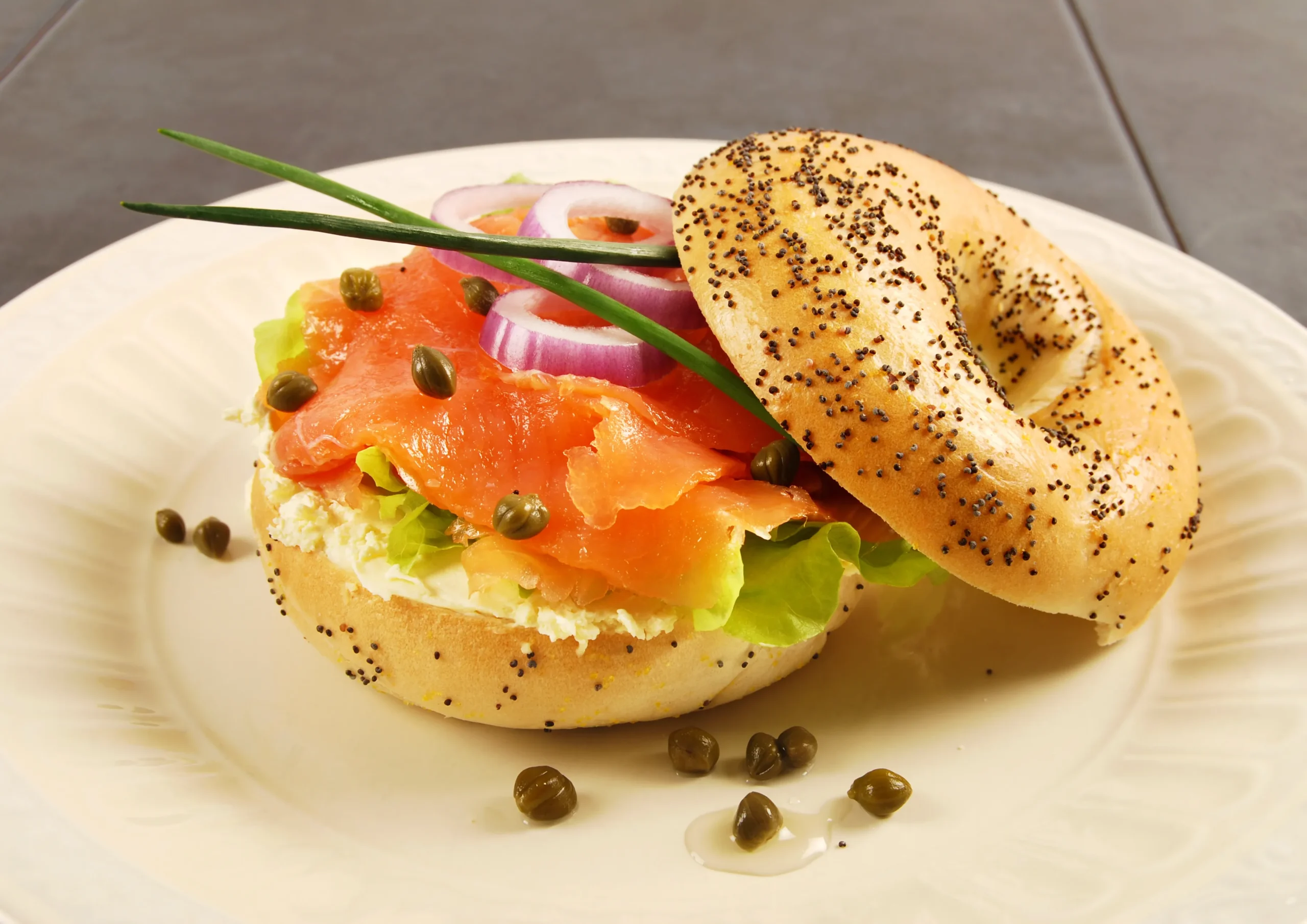 Poppy seed bagel filled with smoked salmon, cream cheese, capers, red onion, and lettuce on a white plate.