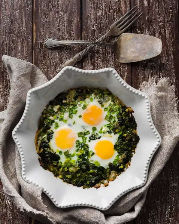 A white ceramic dish filled with spinach and egg scramble, topped with three sunny-side-up eggs and garnished with chopped green onions, placed on a rustic wooden table.