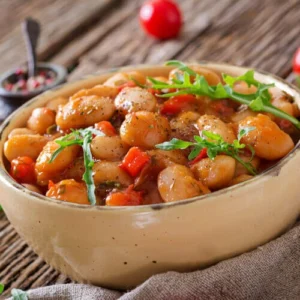 A bowl of hearty bean stew with white beans, diced tomatoes, and fresh arugula garnish, served on a rustic wooden table.