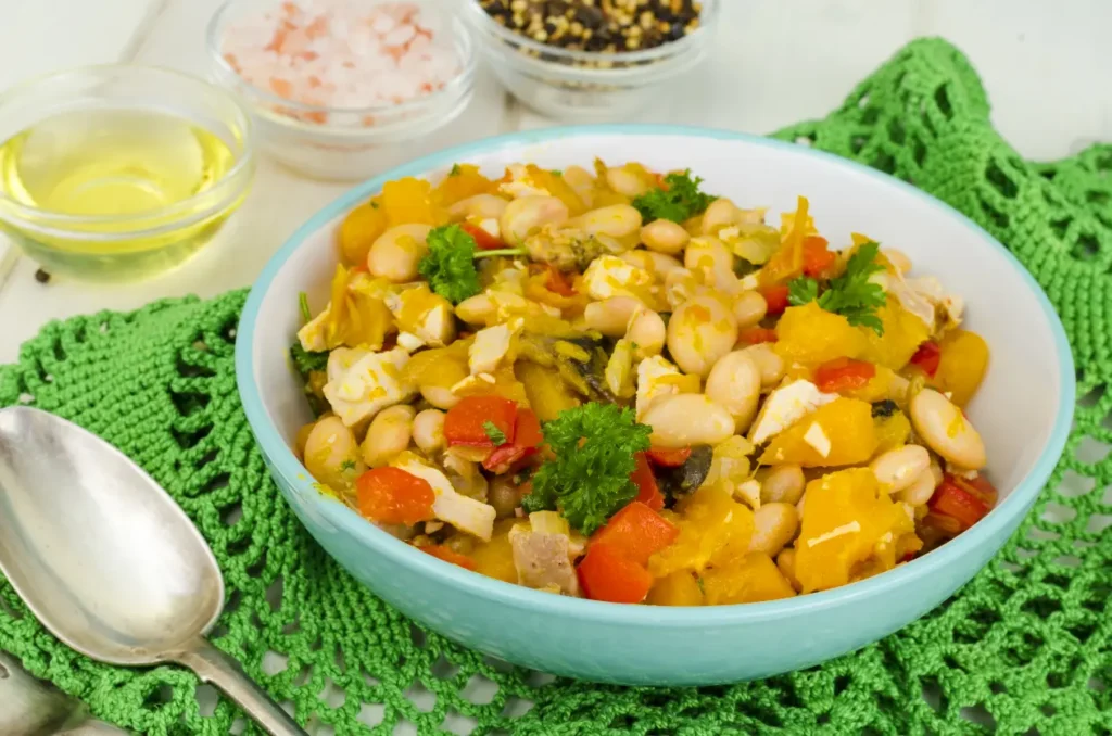 A colorful bowl of bean stew featuring white beans, tender pumpkin chunks, red bell peppers, and fresh parsley, served on a green crocheted mat with spices and oil in the background.