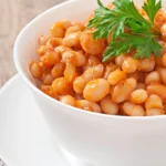 A close-up of a white bowl filled with stewed white beans in a tomato-based sauce, garnished with a sprig of fresh parsley.