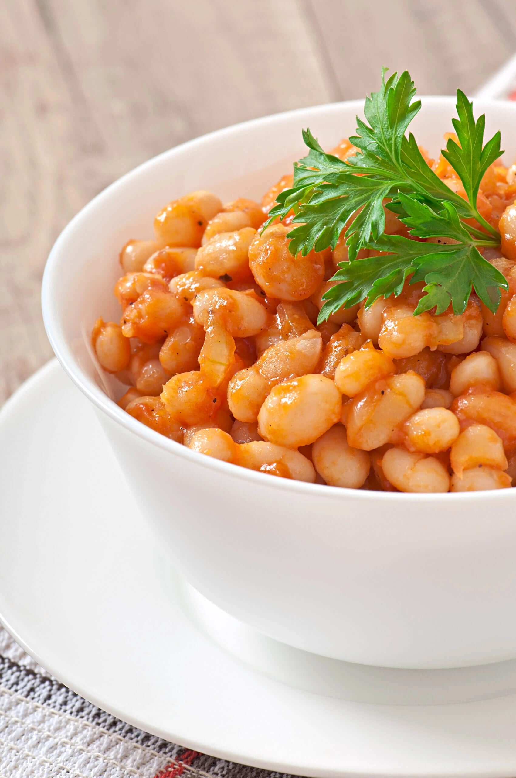 A close-up of a white bowl filled with stewed white beans in a tomato-based sauce, garnished with a sprig of fresh parsley.