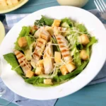 A fresh chicken Caesar salad with grilled chicken, romaine lettuce, croutons, shredded parmesan, and Caesar dressing served in a white bowl on a picnic table.