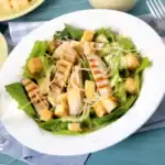 A white bowl filled with chicken Caesar salad, featuring grilled chicken, crisp lettuce, croutons, shredded parmesan, and Caesar dressing on a picnic table.