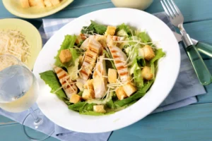 A fresh chicken Caesar salad with grilled chicken, romaine lettuce, croutons, shredded parmesan, and Caesar dressing served in a white bowl on a picnic table.