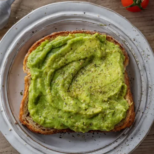 A slice of toasted bread topped with creamy mashed avocado, seasoned with salt and pepper, served on a white plate with avocado, tomatoes, and seasoning ingredients nearby.