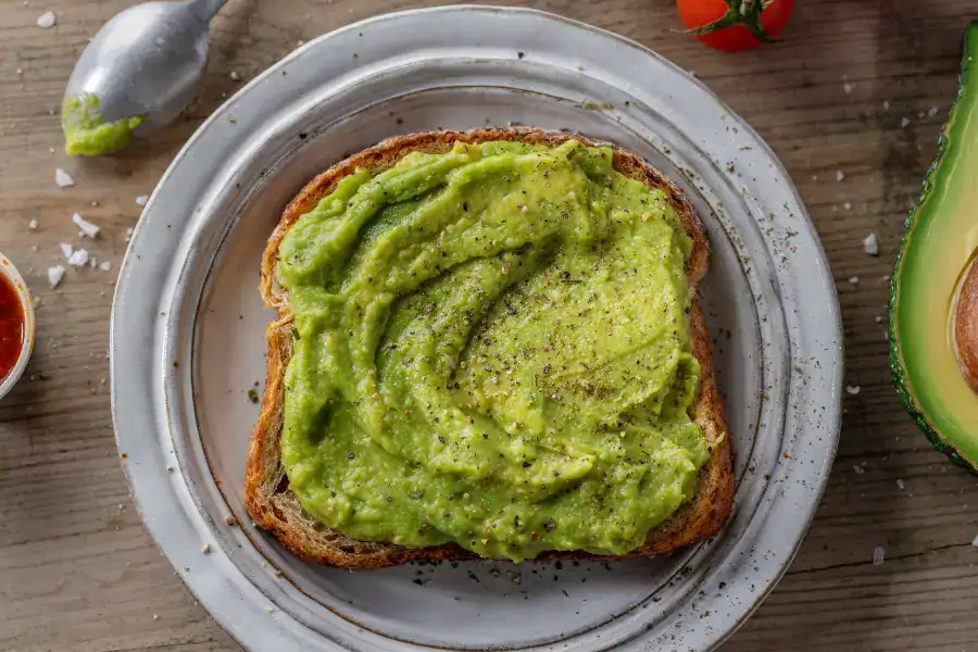 A slice of toasted bread topped with creamy mashed avocado, seasoned with salt and pepper, served on a white plate with avocado, tomatoes, and seasoning ingredients nearby.