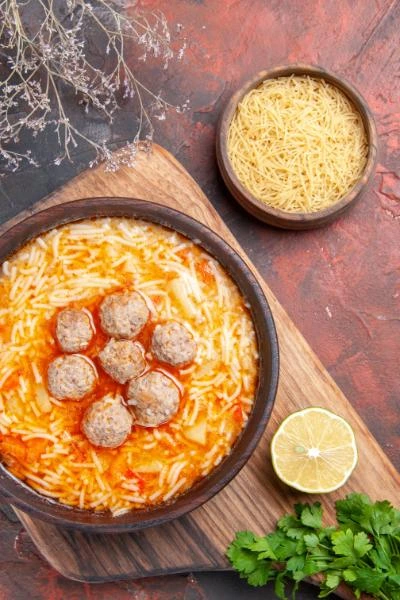 A bowl of pasta meatball soup topped with six meatballs, surrounded by thin noodles in a tomato-based broth, placed on a wooden board with lemon, parsley, and uncooked noodles nearby.