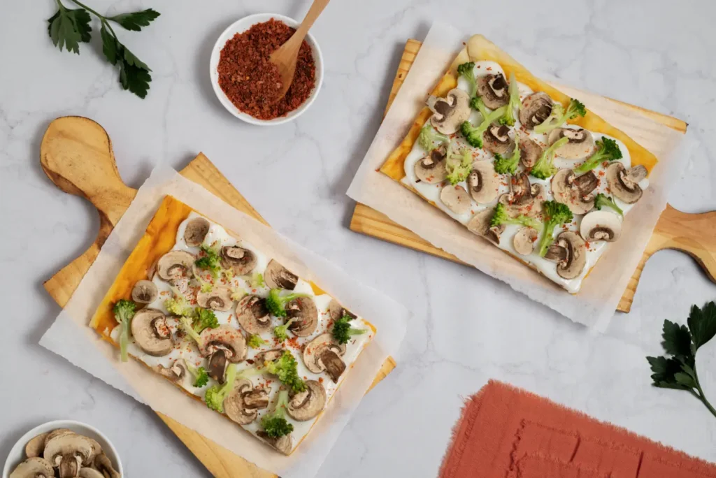 Two rectangular flatbread mushroom pizzas topped with fresh sliced mushrooms, broccoli florets, and a creamy white sauce, served on parchment paper over wooden boards. Two rectangular flatbread mushroom pizzas topped with fresh sliced mushrooms, broccoli florets, and a creamy white sauce, served on parchment paper over wooden boards.