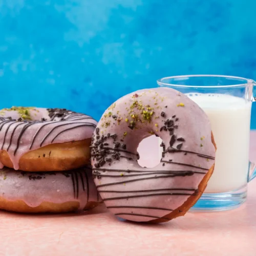 Three pink glazed doughnuts with chocolate drizzles and pistachio toppings next to a glass of milk on a pink table.