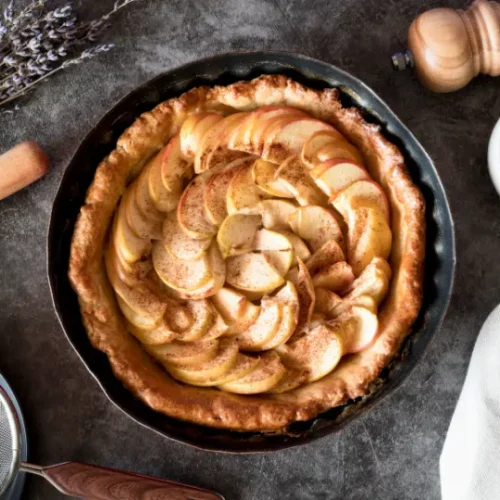 A freshly baked apple pie in a round tray with thinly sliced apples arranged in a spiral, surrounded by baking tools, lavender, and a jug of milk on a dark textured background.