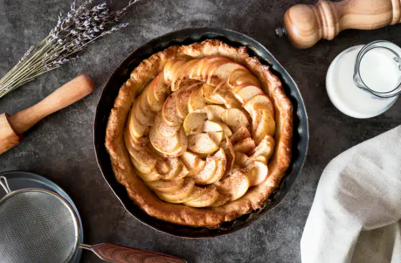 A freshly baked apple pie in a round tray with thinly sliced apples arranged in a spiral, surrounded by baking tools, lavender, and a jug of milk on a dark textured background.