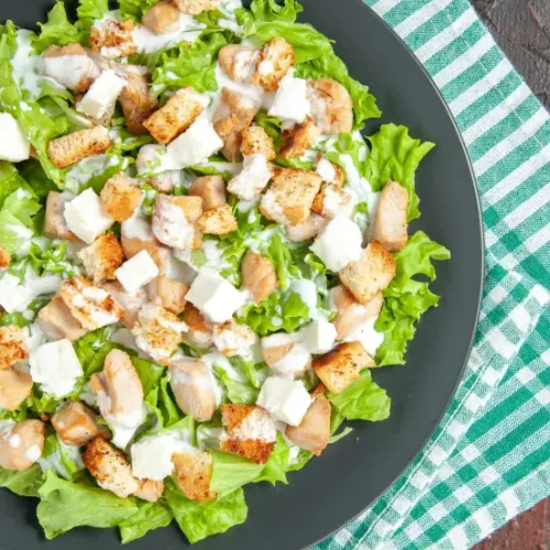 A top-view of Chicken Caesar Salad served on a dark plate, featuring fresh lettuce, grilled chicken chunks, croutons, feta cheese, and creamy dressing. The plate is placed on a green and white checkered tablecloth.