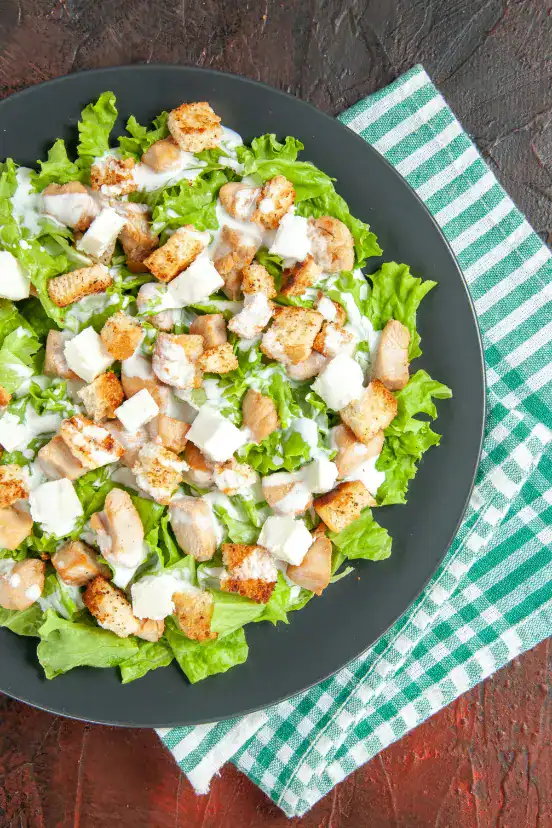 A top-view of Chicken Caesar Salad served on a dark plate, featuring fresh lettuce, grilled chicken chunks, croutons, feta cheese, and creamy dressing. The plate is placed on a green and white checkered tablecloth.