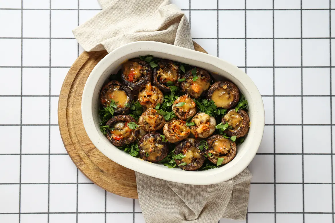 A baking dish filled with stuffed mushrooms topped with melted cheese, fresh parsley, and vibrant filling, placed on a wooden board with a light-colored towel.