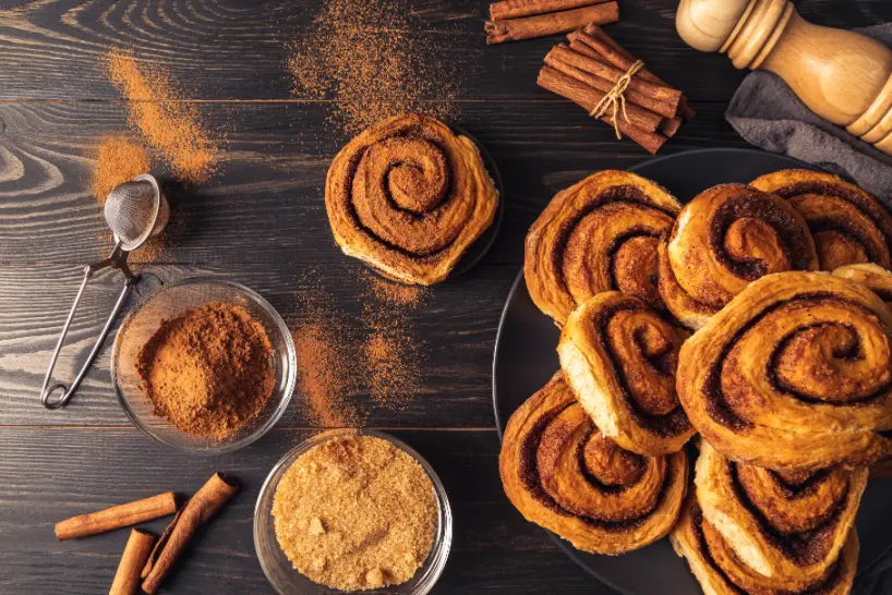 A top view of cinnamon rolls on a black plate surrounded by cinnamon sticks, brown sugar, and powdered cinnamon on a wooden table.