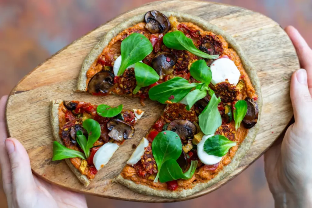 A vibrant vegan mushroom pizza served on a wooden board, topped with fresh greens, mushrooms, tomato sauce, and dollops of plant-based cream.