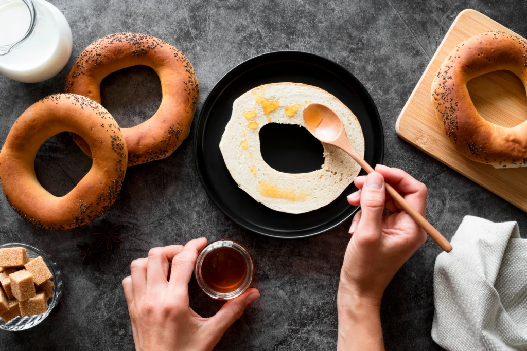 A hand spreading honey on a sliced bagel with a wooden spoon, surrounded by whole bagels and accompaniments on a dark surface.