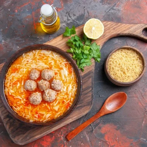 A rustic bowl of pasta meatball soup on a wooden board, surrounded by uncooked noodles, fresh parsley, a halved lemon, and a bottle of olive oil, with a wooden spoon beside it.