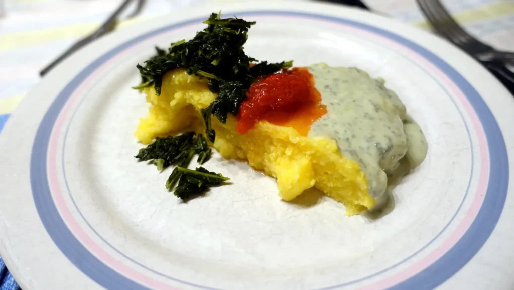 A serving of polenta on a white plate, topped with creamy herb sauce, a dollop of tomato sauce, and sautéed greens.