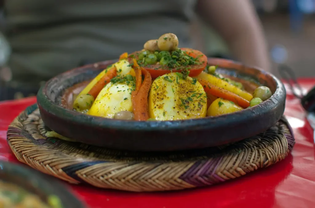 A colorful chicken tagine topped with carrots, potatoes, tomatoes, and green olives, served in a rustic clay dish on a woven mat.